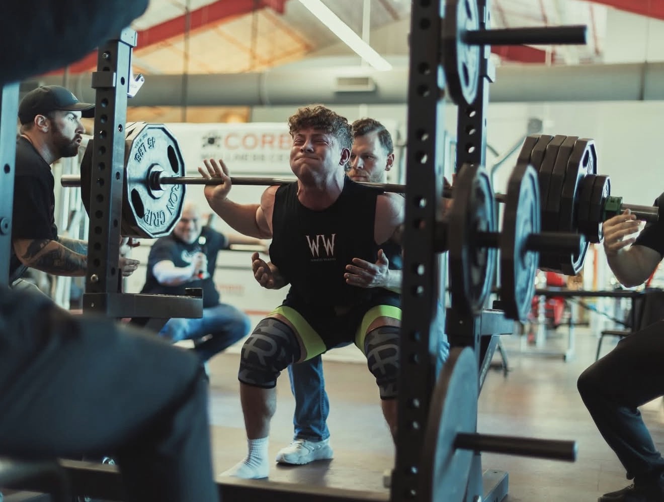 Athlete squatting heavy in a Whitley Weights singlet with spotters